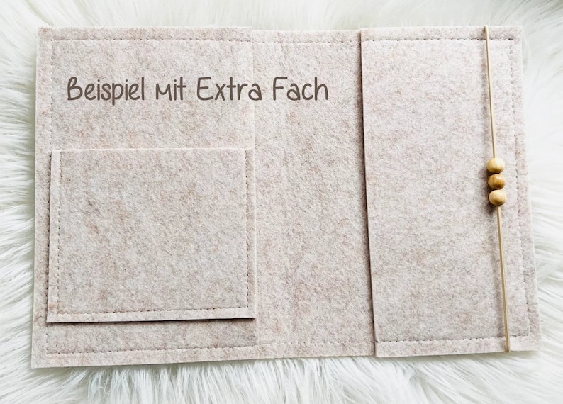 May include: A beige felt organizer with a pocket and a string closure with wooden beads. The text "Beispiel mit Extra Fach" is printed on the felt. The organizer is designed to hold items.