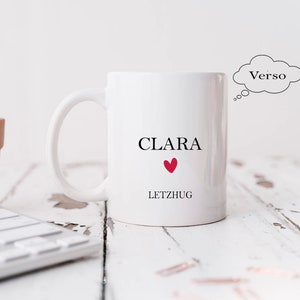 May include: White ceramic mug with the name "CLARA" and the word "LETZHUG" printed on it. A red heart is between the words. The mug is on a white wooden surface.