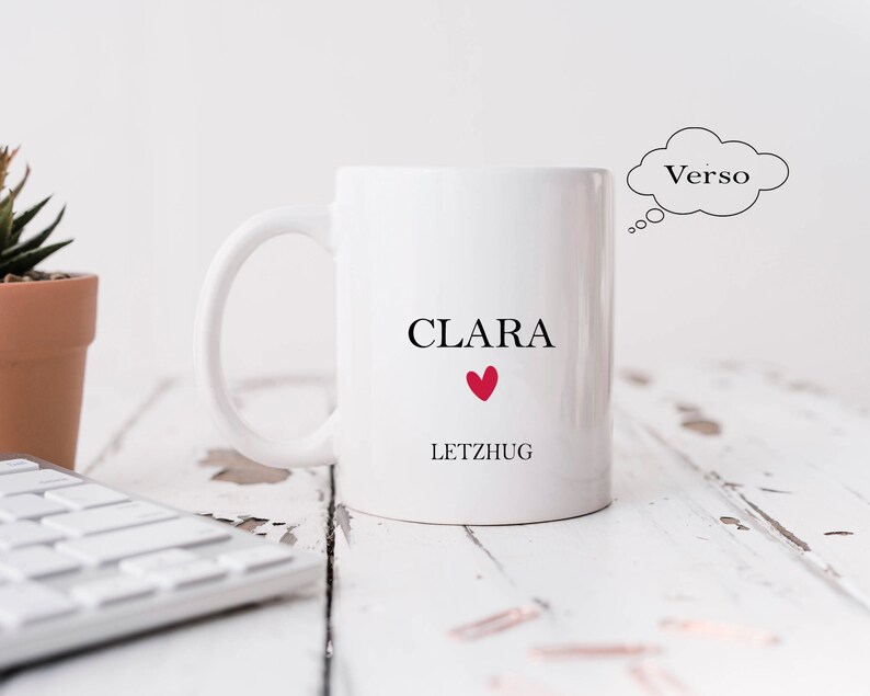 May include: White ceramic mug with the name "CLARA" and the word "LETZHUG" printed on it. A red heart is between the words. The mug is on a white wooden surface.