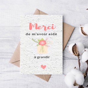 May include: A white card with the text "Merci de m'avoir aid&eacute; &agrave; grandir" in pink and black lettering. A pink flower with green leaves is illustrated in the centre of the card. A pink heart is in the bottom right corner.