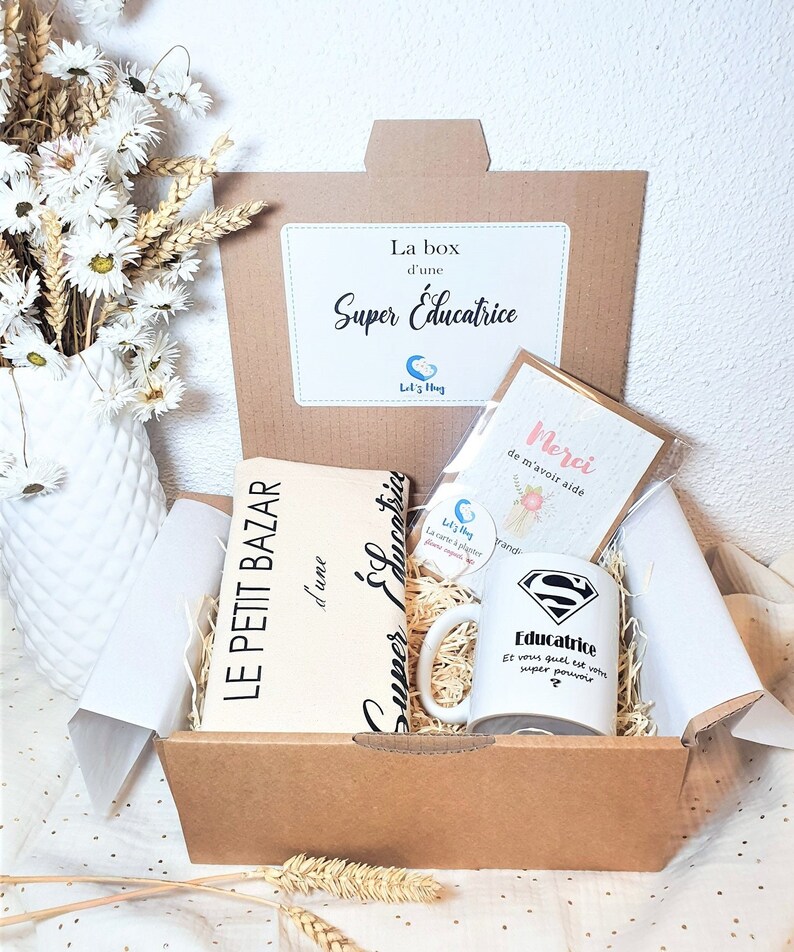 May include: A brown cardboard box with the text "La box d'une Super Educatrice" on the top. The box is open and contains a white mug with the text "Educatrice" and a superhero symbol, a small card with the text "Merci de m'avoir aide", and a notebook with the text "Le Petit Bazar d'une Super Educatrice".