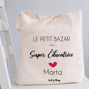 May include: White canvas tote bag with black text that reads "Le Petit Bazar d'une Super Educatrice Marta Let's Hug" with a red heart.