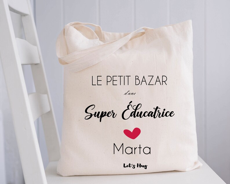 May include: White canvas tote bag with black text that reads "Le Petit Bazar d'une Super Educatrice Marta Let's Hug" with a red heart.