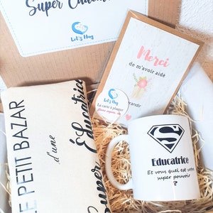 May include: A gift box filled with items for a teacher. The box contains a white mug with a superhero logo and the text "Educatrice Et vous quel est votre super pouvoir?"  A white tote bag with the text "Super Educatrice" and a card with the text "Merci de m'avoir aid&eacute;".