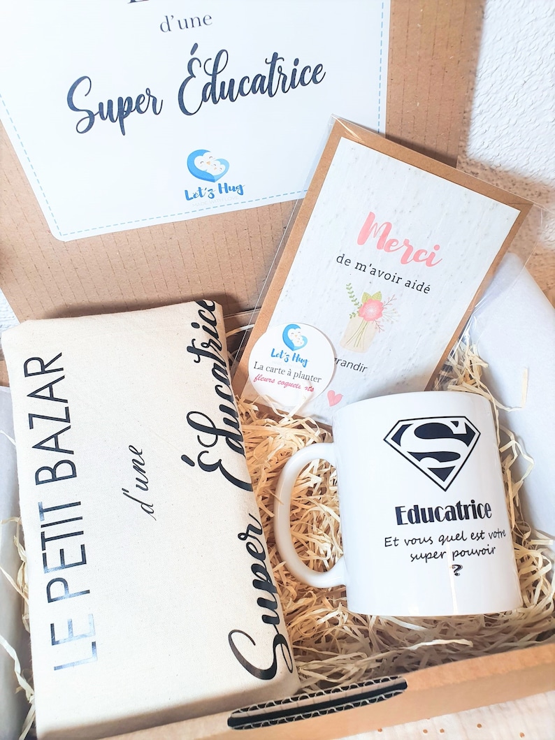 May include: A gift box filled with items for a teacher. The box contains a white mug with a superhero logo and the text "Educatrice Et vous quel est votre super pouvoir?"  A white tote bag with the text "Super Educatrice" and a card with the text "Merci de m'avoir aid&eacute;".