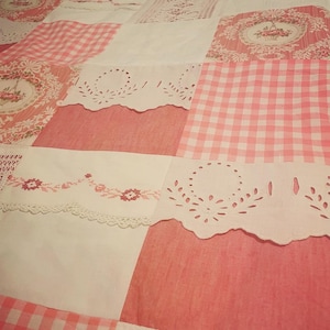 Könnte beinhalten: Eine Patchwork-Decke mit einem rosa-weißen Karomuster, weißem Spitzenbesatz und einem rosafarbenen Blumendesign. Die Decke ist aus verschiedenen Stoffen gefertigt und hat einen handgefertigten Charakter. Das Etikett auf der Decke lautet "Dormischen Design."