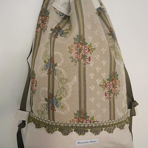 Könnte beinhalten: Ein grüner und weiß gestreifter Rucksack mit Kordelzug, floralem Muster und grünem Fransenbesatz. Die Tasche hat ein weißes Etikett mit dem Text "Dornseifen Design".