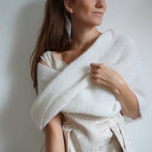 Könnte beinhalten: Ein weißer, gestrickter Schal, der über einem Kleid drapiert ist. Der Schal ist aus einem weichen, strukturierten Garn gefertigt. Das Kleid hat eine helle, schimmernde Farbe. Der Gesamteindruck ist elegant und für besondere Anlässe geeignet.