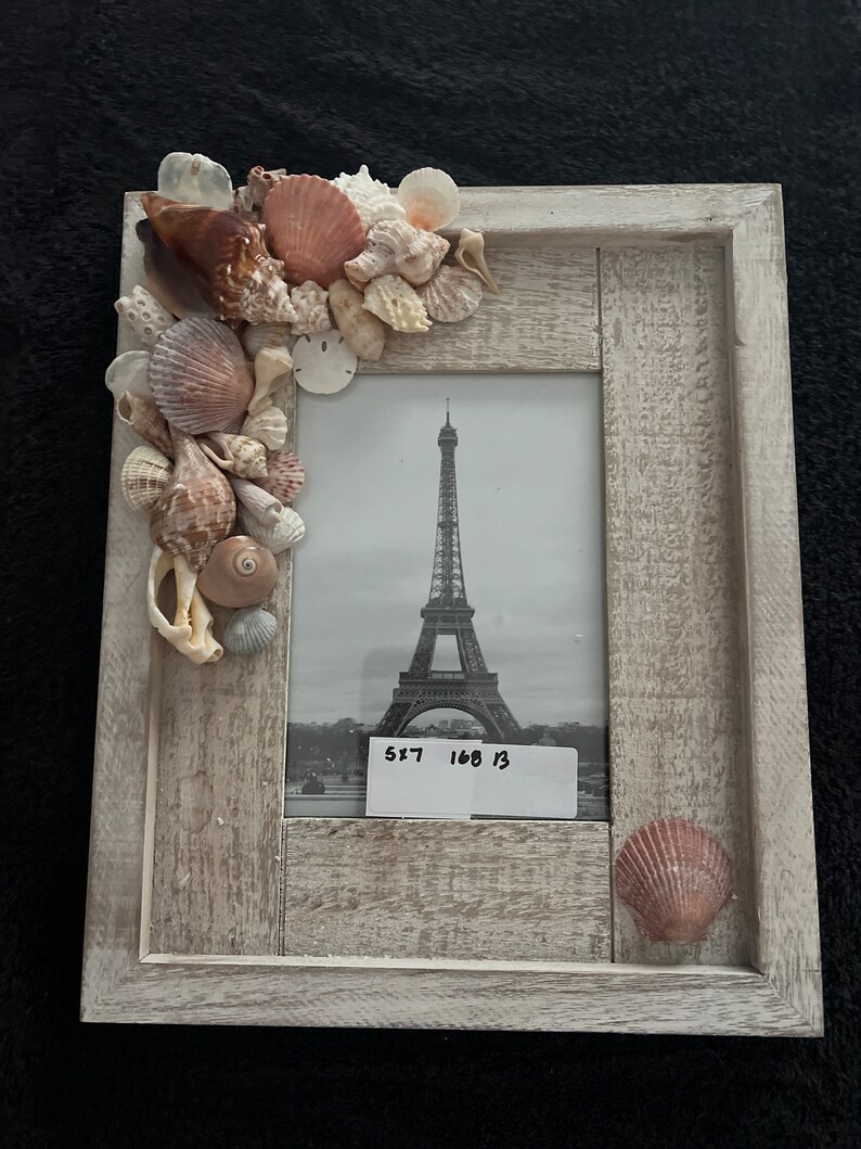 May include: A white wooden picture frame with a black and white photo of the Eiffel Tower. The frame is decorated with seashells in shades of brown, white, and pink. The text "5x7 168 13" is written on a white label in the bottom right corner of the photo.