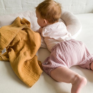 May include: A baby wearing a white tank top and pink ruffled bloomers is lying on a white waffle-textured pillow. A mustard yellow muslin blanket is lying on the bed next to the baby.