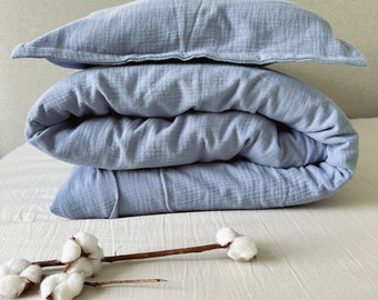 Puede incluir: Un juego de funda nórdica y funda de almohada de algodón azul claro. La funda nórdica está doblada y la funda de almohada está encima. El juego está sobre una cama blanca.