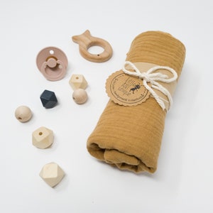 May include: A mustard yellow muslin swaddle blanket rolled up with a brown tag that says "Little Ant Unique" and a white string tied around it.  There are wooden beads, a wooden fish toy, and a pink silicone pacifier on a white background.