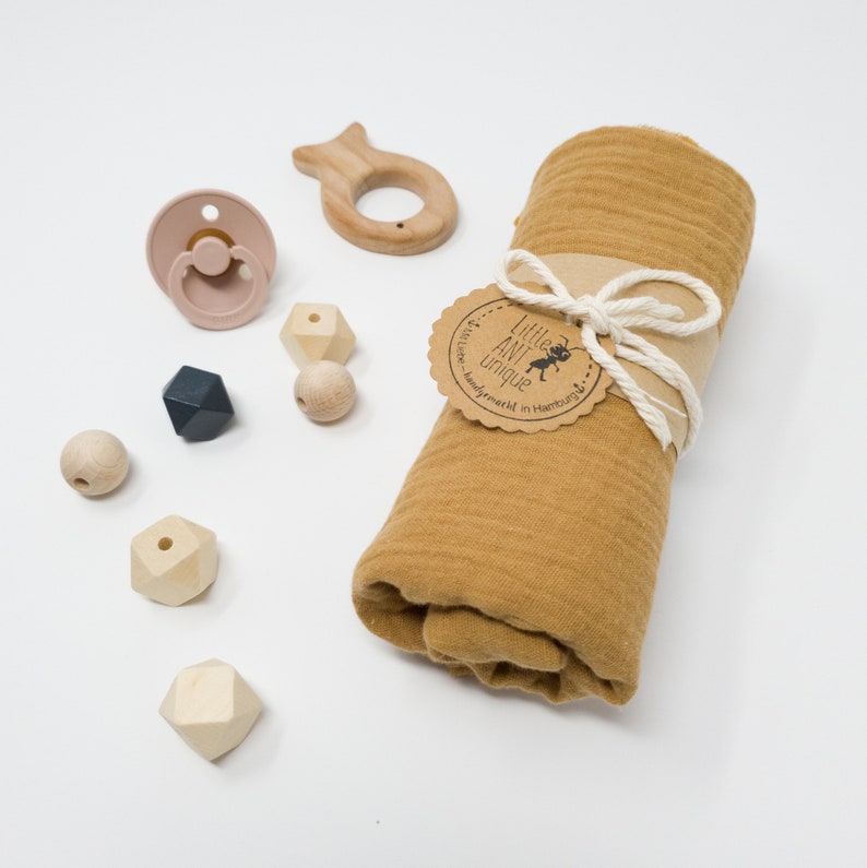 May include: A mustard yellow muslin swaddle blanket rolled up with a brown tag that says "Little Ant Unique" and a white string tied around it.  There are wooden beads, a wooden fish toy, and a pink silicone pacifier on a white background.