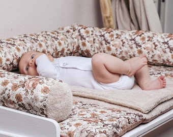 May include: A baby lying on a floral patterned baby nest with a white onesie. The nest is made of soft fabric and has a brown and white floral pattern.