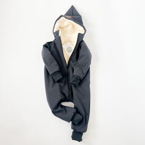 Könnte beinhalten: Schwarzer Kapuzen-Winteroverall mit Fleece-Futter. Der Overall hat einen Reißverschluss und eine Kapuze mit spitzer Spitze.