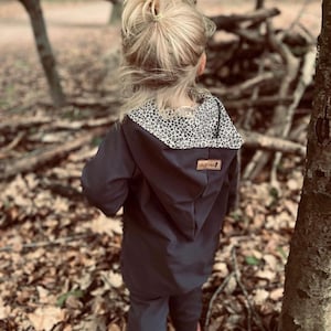 Könnte beinhalten: Eine schwarze Kapuzenjacke mit einem braunen und weißen Tierdruckfutter. Die Jacke hat einen Reißverschluss und ein kleines braunes Etikett mit dem Text "Little Monkey" auf der Vorderseite.