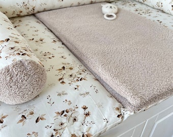 May include: A white changing pad cover with a brown floral pattern, featuring matching side bolsters with brown textured ends. A rectangular beige textured mat rests on the pad, accompanied by a small white circular rattle with a cat face.
