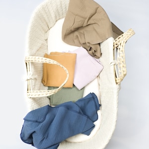 May include: A white wicker basket with a blue, green, brown, and pink muslin swaddles inside. The swaddles are folded and arranged in the basket.