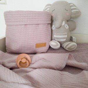 May include: A pink waffle weave storage basket with a brown label, a gray knitted elephant toy, and a pink muslin blanket with a beige pacifier.