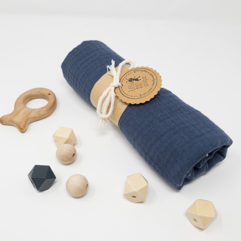 May include: A blue muslin swaddle blanket rolled up and tied with twine. The blanket has a brown tag with the text "Curiosity is Natural" and a small illustration of an ant. The image also includes a wooden fish-shaped teether and wooden beads in various shapes.