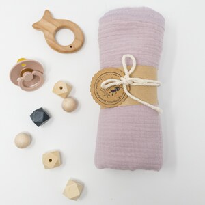 May include: A pink muslin swaddle blanket with a brown tag that says "handmade with love in Hamburg" and a small ant logo. The blanket is tied with a natural twine bow.  The image also includes a wooden fish teether, a pink silicone pacifier, and wooden geometric shapes.