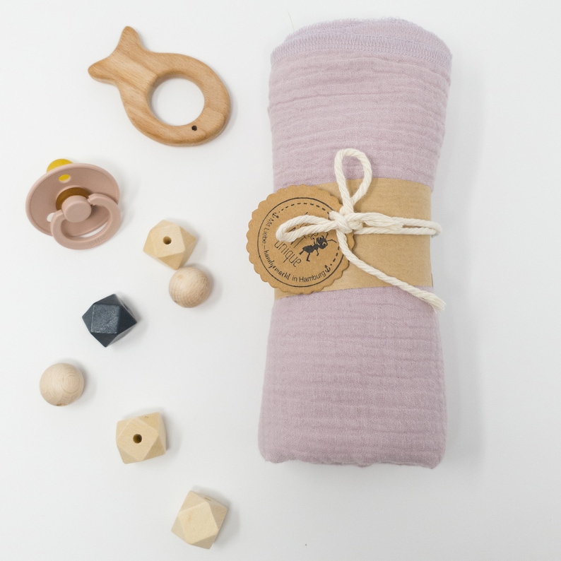 May include: A pink muslin swaddle blanket with a brown tag that says "handmade with love in Hamburg" and a small ant logo. The blanket is tied with a natural twine bow.  The image also includes a wooden fish teether, a pink silicone pacifier, and wooden geometric shapes.