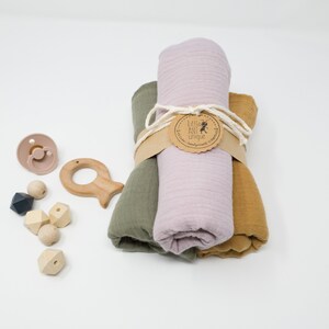 May include: A set of three muslin swaddles in green, pink, and yellow, rolled up and tied with twine. The swaddles are labeled with a brown tag that reads "Little Ant Unique Handmade Treasures". The set also includes a wooden teether shaped like a fish, a pacifier, and wooden beads.