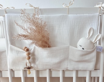 May include: A white crib organizer with two pockets, attached to the crib with ribbons. One pocket holds a stuffed bunny, the other holds dried plants. A wooden pacifier clip hangs from the organizer.