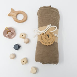 May include: A brown muslin swaddle blanket with a brown paper tag that says "Little Ant Unique" with a small ant illustration. The blanket is rolled up and tied with twine. There are several wooden toys scattered around the blanket, including a wooden fish, a pacifier, and a few wooden blocks.