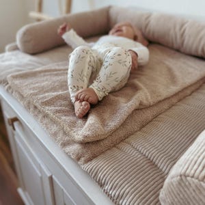Könnte beinhalten: Eine beigefarbene Wickelauflage aus Cord mit erhöhten Seitenpolstern und einer weichen, strukturierten Oberfläche. Die Auflage verfügt über einen abnehmbaren, plüschigen Bezug in Beige und bietet eine gepolsterte Fläche zum Wickeln.