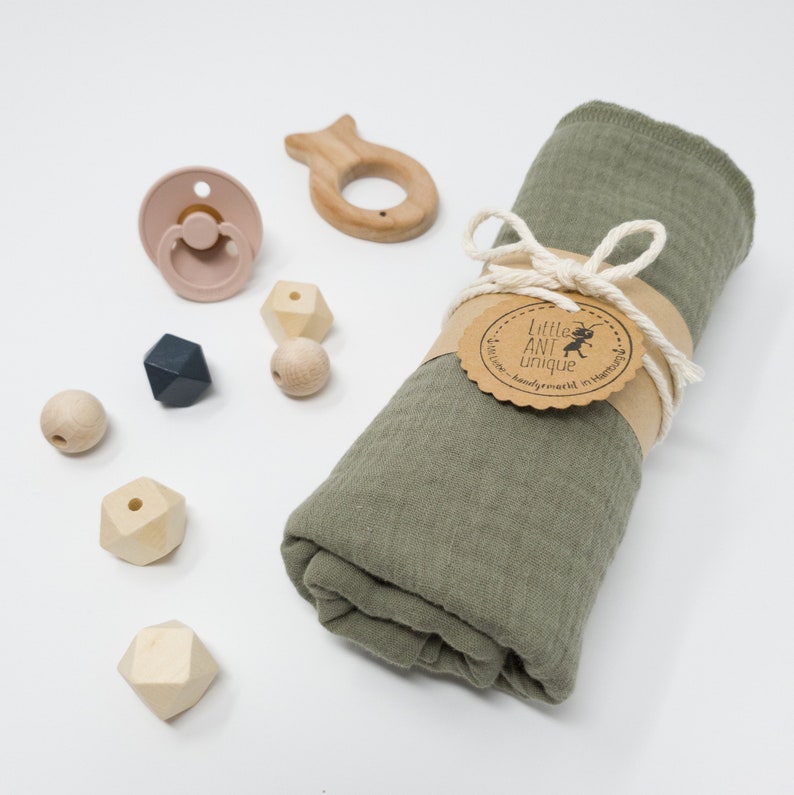May include: A green muslin swaddle blanket with a brown tag that reads "Little Ant Unique - handmade in Hamburg" is rolled up and tied with twine. The blanket is surrounded by wooden toys, including a fish-shaped teether, a pacifier, and geometric-shaped beads.