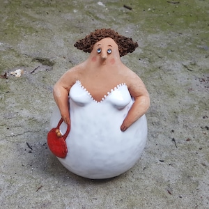 Peut inclure: Une figurine en céramique d'une femme portant une robe blanche et tenant un sac à main rouge. La femme a les cheveux bruns et les yeux bleus.