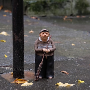 以下が含まれることがあります： 茶色の横縞のセーター、濃い色のズボン、帽子をかぶり、ほうきを持っている男性の陶器製フィギュア。 濡れた表面に落ち葉があり、黒い棒の隣に立っています。