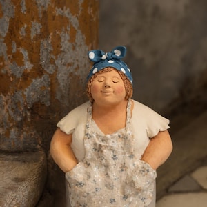 Könnte beinhalten: Eine Keramikfigur einer Person mit geschlossenen Augen, bekleidet mit einem weißen kurzärmeligen Oberteil, einer beigen geblümten Schürze mit Taschen und einem blauen Kopftuch mit weißen Punkten. Die Figur hat eine glatte, zylindrische Basis.