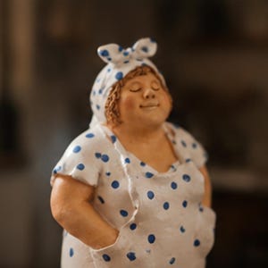 Könnte beinhalten: Eine Keramikfigur einer Person mit geschlossenen Augen und einem sanften Lächeln, die ein weißes Kleid und ein passendes Kopftuch trägt, beide mit blauen Punkten verziert. Die Figur hat die Hände in den Taschen des Kleides.