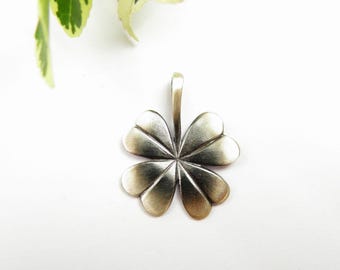 May include: A silver-toned clover pendant with four heart-shaped leaves. The pendant has a loop at the top for a chain. The background is white with a sprig of green and white leaves in the upper left corner.