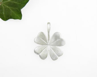 Op de afbeelding: Zilverkleurige vierbladig klaverblad hanger met een lus voor een ketting. De hanger staat tegen een witte achtergrond met een groen blad in de linkerbovenhoek. De blaadjes van de klaver hebben een textuur.
