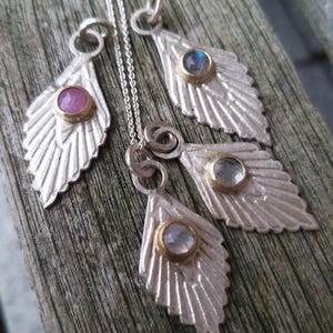 Könnte beinhalten: Drei silberne, blattartige Anhänger mit goldenen Akzenten und verschiedenen farbigen Edelsteinen. Die Edelsteine sind rosa, blau und weiß.