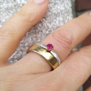 Könnte beinhalten: Ein zweifarbiger Ring mit einem goldenen Band und einem silbernen Band. Der Ring hat einen einzigen, kleinen, leuchtend pinkfarbenen Edelstein in der Mitte.