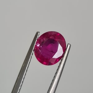 May include: A round, deep red gemstone held by tweezers.