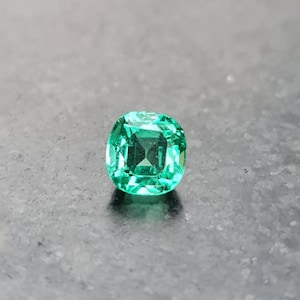 Puede incluir: Una gema de esmeralda verde vibrante con un corte de coj&iacute;n. La gema est&aacute; colocada sobre un fondo gris.