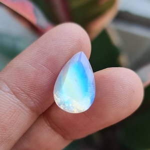 May include: A teardrop-shaped iridescent gemstone with a pearlescent sheen. The gemstone is held between two fingers.