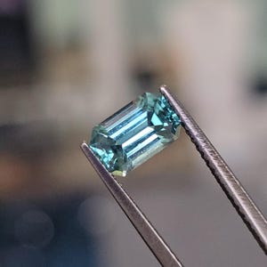 May include: A light blue emerald cut gemstone held by tweezers. The gemstone has a slight green hue.