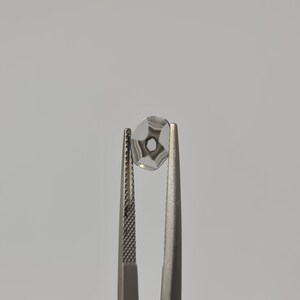 May include: A clear glass lens with a star-shaped pattern is held by tweezers. The lens has a small hole in the center.