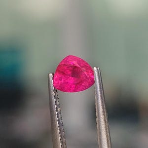 May include: A vibrant pink gemstone with a triangular shape, held in tweezers.