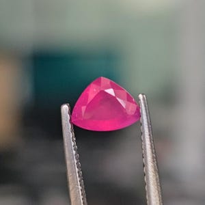 May include: A triangular, pink gemstone held in tweezers. The gemstone is faceted and has a bright, vibrant colour.