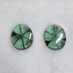 May include: Two oval-shaped, translucent green gemstones with a starburst pattern of dark inclusions. The gemstones are set against a white background, showcasing their clarity and colour.