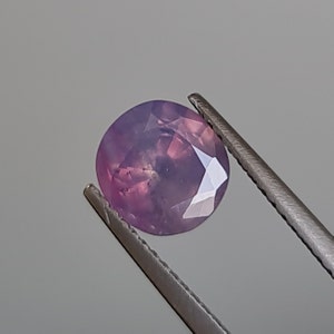 May include: A round, faceted, purple gemstone with a light pink hue. The gemstone is held in tweezers.