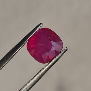 May include: A vibrant red cushion-cut ruby gemstone held by tweezers.
