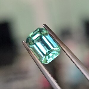 May include: A vibrant green emerald cut gemstone held in tweezers. The gemstone has a brilliant, faceted cut and a translucent appearance.